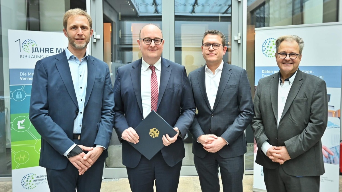 Vier Männer in Anzügen stehen nebeneinander und lächeln in die Kamera. Im Hintergrund sind Roll-ups der Medizininformatik-Initiative zu erkennen.