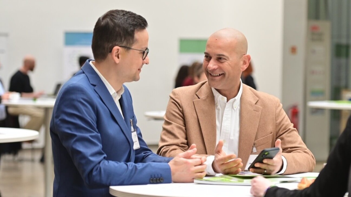 Zwei Männer im Gespräch an einem Tisch, einer hält ein Smartphone und beide lächeln.