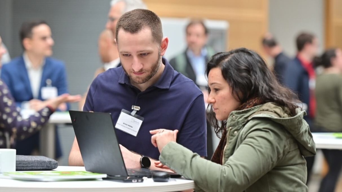 Ein Mann und eine Frau gucken zusammen auf einen Laptop-Bildschirm. Im Hintergrund sind andere Menschen zu sehen, die sich unterhalten.
