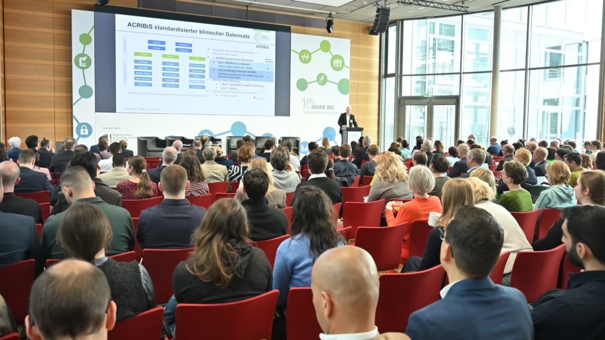 Ein großes Publikum hört einem Vortrag zu; im Hintergrund sind eine PowerPoint und ein Mann vor einem Rednerpult zu erkennen.