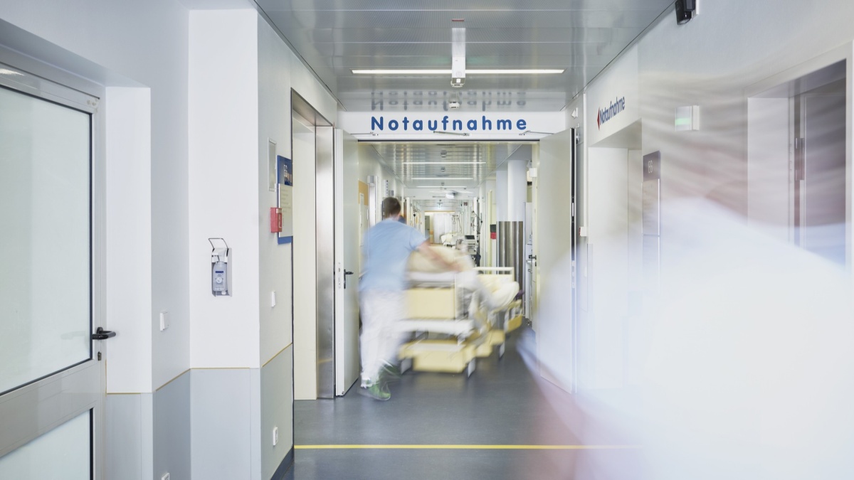 Heller Krankenhausflur mit der Beschilderung ‚Notaufnahme‘ im Hintergrund. Eine medizinische Fachkraft schiebt unscharf ein Krankenbett durch den Gang, wodurch der Eindruck von Eile und Bewegung entsteht.
