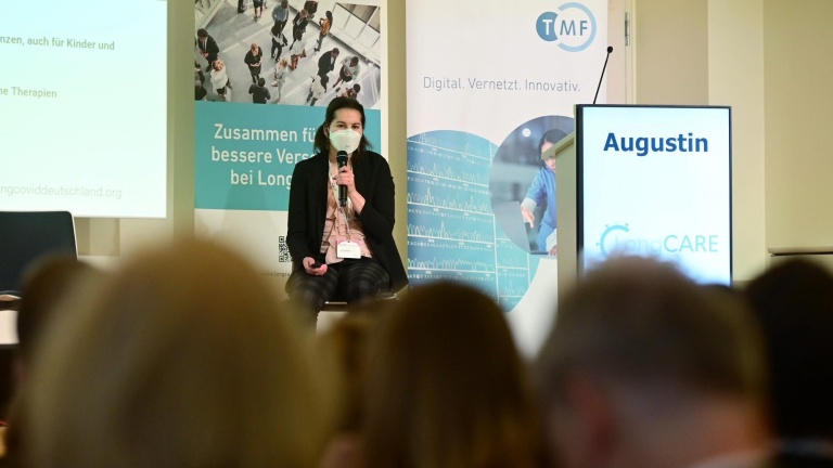 Eine Frau mit einer Atemschutzmaske und einem Mikrofon sitzt vor einem großen Publikum. Hinter ihr sind Roll-ups, unter anderem mit dem Logo der TMF e.V.