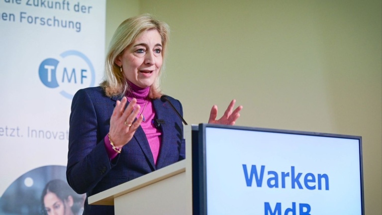 Eine Frau steht an einem Rednerpult und spricht mit erhobenen Händen zu einem Publikum. Sie trägt ein Sakko über einem farbigen Rollkragenpullover. Auf dem Pult ist ein Bildschirm mit dem Text ‚Warken MdB‘ zu sehen. Im Hintergrund befindet sich ein Banner mit dem Logo ‚TMF‘.