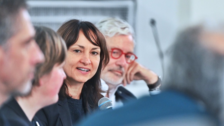 Eine Frau spricht in ein Mikrofon. Neben ihr sitzen weitere Personen und hören zu.