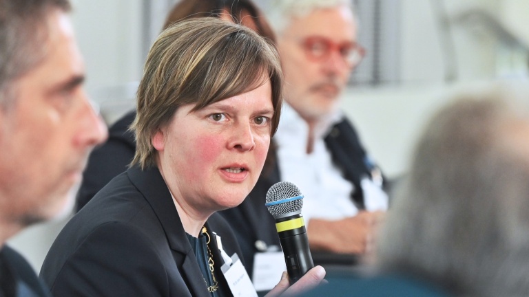 Eine Frau spricht in ein Mikrofon. Neben ihr sitzen weitere Teilnehmende der Paneldiskussion und hören zu.