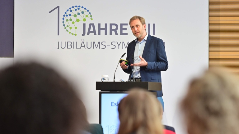 Ein Mann im blauen Anzug steht an einem Rednerpult und hält einen Vortrag vor Publikum; im Hintergrund ist ein Banner mit der Aufschrift „10 Jahre MII Jubiläums-Symposium“ zu sehen.