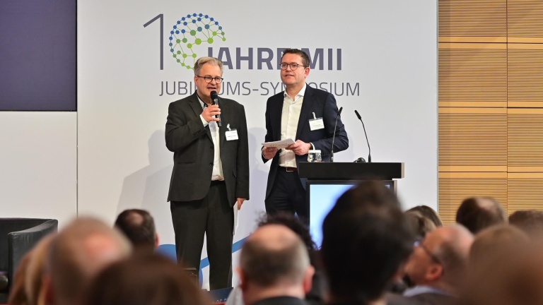 Zwei Männer in Anzügen vor einem Rednerpult; im Hintergrund ist ein Banner mit der Aufschrift „10 Jahre MII Jubiläums-Symposium“ zu sehen.