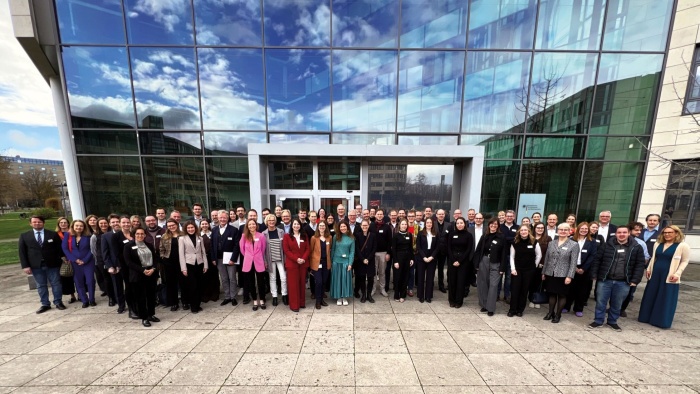 Großes Gruppenfoto vor dem modernen Glasgebäude des BfArM in Bonn. Die Personen stehen in mehreren Reihen vor dem Haupteingang, lächeln in die Kamera und tragen überwiegend Business-Kleidung. Die Glasfassade spiegelt den blauen Himmel mit Wolken.