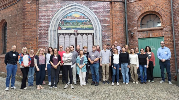 Gruppenfoto von rund 30 Personen, die vor einer historischen Backsteinfassade mit einem großen gotischen Bogenportal stehen. Im Bogen befindet sich eine kunstvoll gestaltete Tafel mit einem farbigen Mosaik und Inschriften. Die Gruppe blickt überwiegend in die Kamera und ist in mehreren Reihen angeordnet. Die Teilnehmenden tragen Alltagskleidung, einige haben Namensschilder um den Hals. Rechts ist eine grüne Tür in die Backsteinfassade eingelassen, im Vordergrund befindet sich ein gepflasterter Platz.