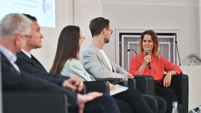 Eine Frau in einer korallfarbenen Bluse spricht bei einer Podiumsdiskussion in ein Mikrofon. Neben ihr sitzen weitere Teilnehmende in Sesseln auf einer Bühne, während im Hintergrund eine Präsentationsfolie zu sehen ist.