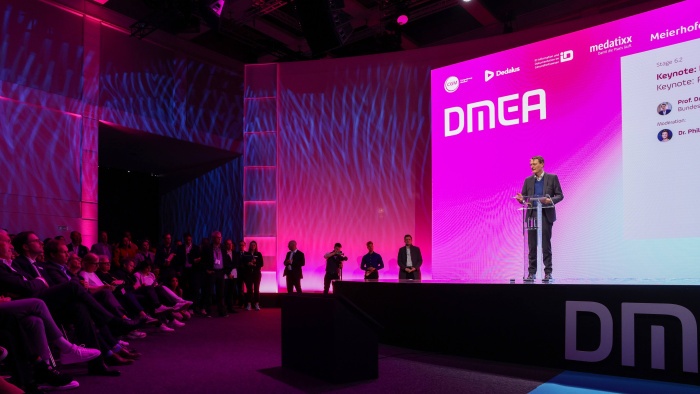 Ein Redner steht auf einer Bühne mit großem pink beleuchtetem Hintergrund und dem Schriftzug „DMEA“. Er spricht an einem transparenten Rednerpult vor einem sitzenden Publikum auf der linken Seite.