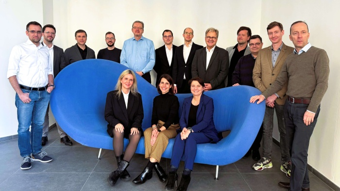 Mehrere Personen stehen hinter einer modernen, blauen Couch, auf der drei Personen sitzen. Sie lächeln für das Gruppenfoto in die Kamera.