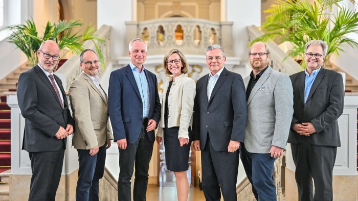 Gruppenfoto des neu gewählten TMF-Vorstands: Sieben Personen stehen nebeneinander in einem repräsentativen Treppenhaus und blicken in die Kamera. Im Hintergrund sind eine breite Marmortreppe mit Balustrade sowie große grüne Pflanzen zu sehen.