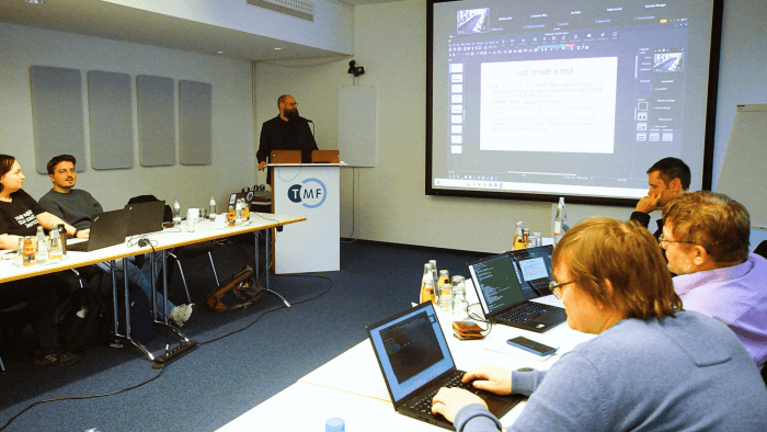 Ein Mann steht vor einem TMF-Rednerpult mit Mikrofon und sieht zu einer PowerPoint neben sich. Vor ihm sitzen an mehreren Tischen Personen mit Laptops.