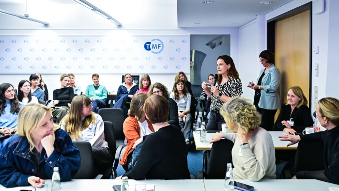 Eine Person steht in der Mitte eines TMF-Veranstaltungsraums und spricht mit einem Handmikrofon, während die übrigen Teilnehmenden des Kick-offs aufmerksam zuhören. Im Hintergrund ist eine Wand mit TMF-Logos zu sehen.