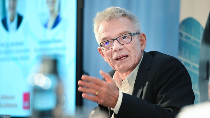 Prof. Josef Hecken mit Anzug und Brille. Er gestikuliert mit einer Hand und vor ihm steht ein Mikrofon. Im Hintergrund ist ein unscharfer Bildschirm mit einer PowerPoint-Präsentation zu erkennen.