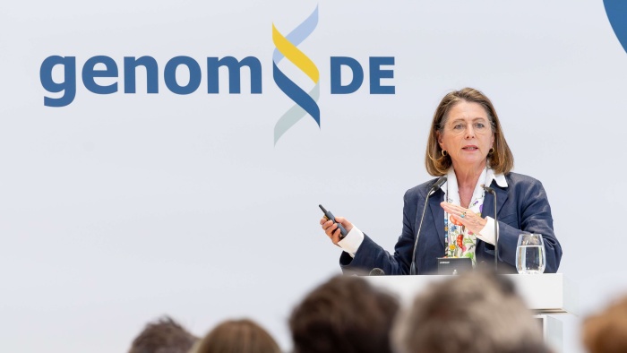 Eine Frau mit schulterlangen Haaren steht hinter einem Rednerpult. Im Hintergrund ist das Logo von genomDE zu sehen.