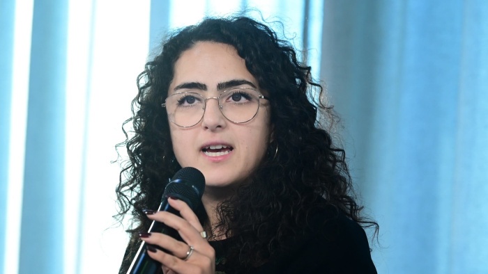 Dr. Nilofar Badra-Azar mit dunklem gelockten Haar und Brille. Sie spricht in ein Mikrofon. Der Hintergrund besteht aus blauen Vorhängen.