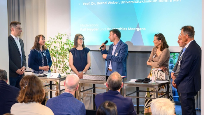 Sechs Personen stehen vor Publikum im Rahmen einer Podiumsdiskussion. Ein Mann in der Mitte spricht in ein Mikrofon. Im Hintergrund wird eine PowerPoint ausgestrahlt.