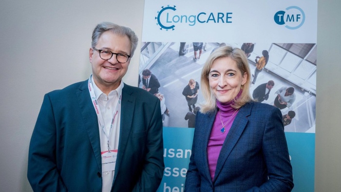 Zwei lächelnde Personen stehen nebeneinander vor einem Roll-up-Banner mit den Logos ‚LongCARE‘ und ‚TMF‘. Beide tragen Businesskleidung; die Person links trägt eine Brille und ein Namensband, die Person rechts ein Sakko über einem farbigen Rollkragenpullover.