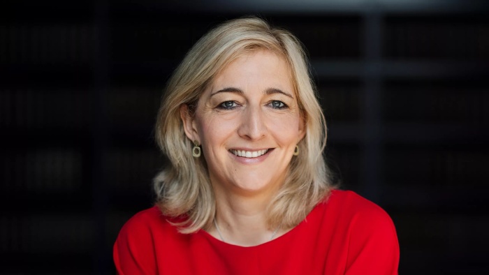 Porträt einer lächelnden Frau mit schulterlangem blondem Haar, die ein rotes Oberteil trägt. Der Hintergrund ist dunkel.