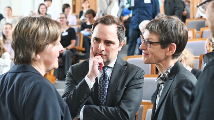 Mehrere Personen in formeller Geschäftskleidung stehen in einem Hörsaal zusammen und führen ein Gespräch. Im Vordergrund diskutieren zwei Frauen mit einem Mann im Anzug, während im Hintergrund weitere Teilnehmende einer Veranstaltung zu sehen sind.
