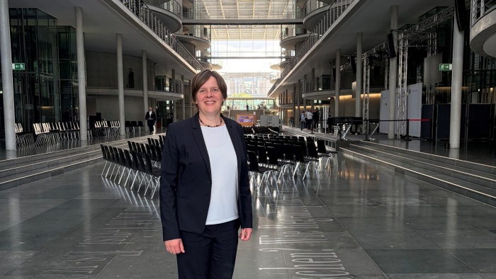 Eine lächelnde Frau im Business-Look steht in einem modernen Gebäude mit zahlreichen Sitzreihen hinter sich, die für eine Veranstaltung platziert wurden.