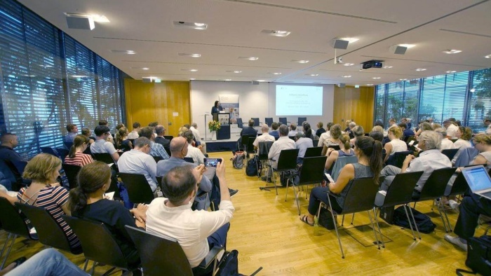 Participants seated in a conference room listen to a speaker at the podium. A presentation is displayed on a screen, and the event appears professional and well attended.