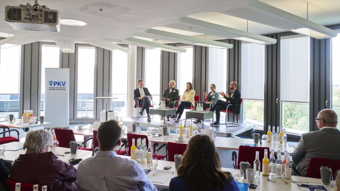 Podiumsdiskussion auf einer Konferenz des PKV-Verbands: Fünf Personen sitzen auf einer Bühne vor großen Fenstern und diskutieren vor dem Publikum. Im Vordergrund stehen Konferenztische mit Getränken, Notizmaterialien und Teilnehmenden.