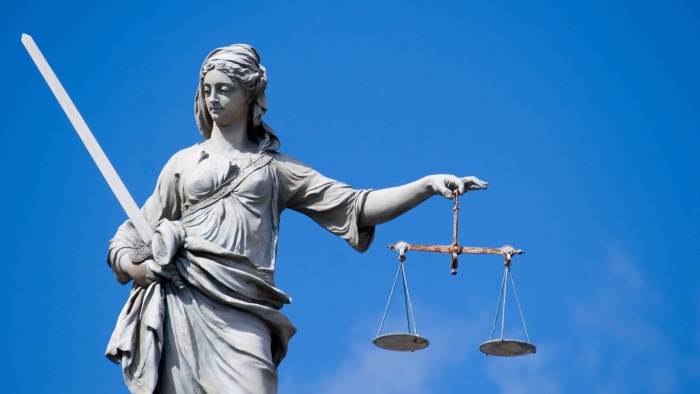 Steinerne Justitia-Statue mit Schwert in der linken Hand und Waage in der rechten, vor blauem Himmel.