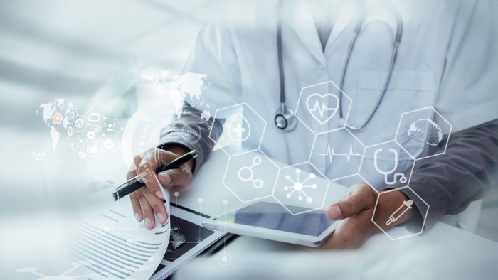 Ärztliche Fachkraft in weißem Kittel mit Stethoskop arbeitet an einem Tablet und hält einen Stift in der Hand. Über dem Bild liegen transparente digitale Gesundheitssymbole wie Herzfrequenz, Gehirn, Spritze und medizinische Vernetzung.