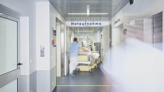 Heller Krankenhausflur mit der Beschilderung ‚Notaufnahme‘ im Hintergrund. Eine medizinische Fachkraft schiebt unscharf ein Krankenbett durch den Gang, wodurch der Eindruck von Eile und Bewegung entsteht.