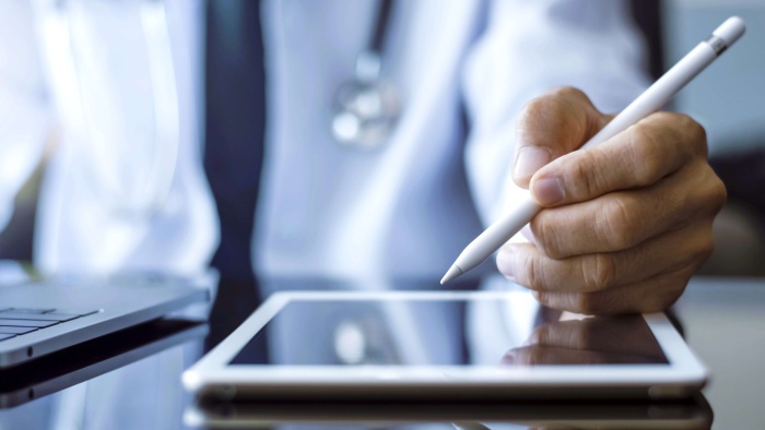Nahaufnahme einer Person in weißem Kittel mit Stethoskop, die mit einem digitalen Stift auf einem Tablet schreibt; daneben steht ein Laptop.