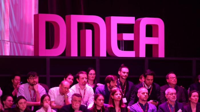 Große pink beleuchtete „DMEA“-Buchstaben auf einer Bühne; davor sitzt ein Publikum aus Frauen und Männern mit Namensschildern und blickt aufmerksam nach vorn.